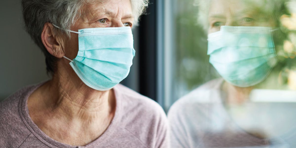 An older woman wearing a sanitation mask looking out a window